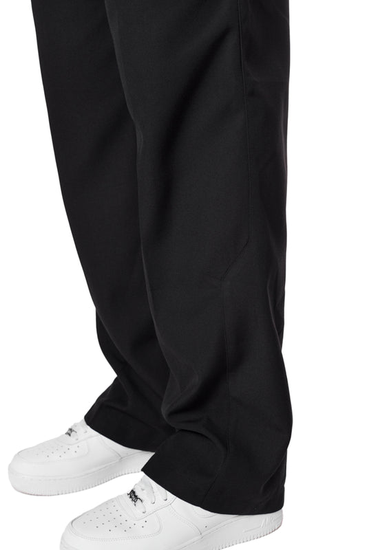Beine in CC Dress Pants Regular Black mit französischen Taschen, asymmetrischem Eingriff, Bundfalten und OLAKALA Logo über der rechten Gesäßtasche, kombiniert mit weißen Schuhen.
