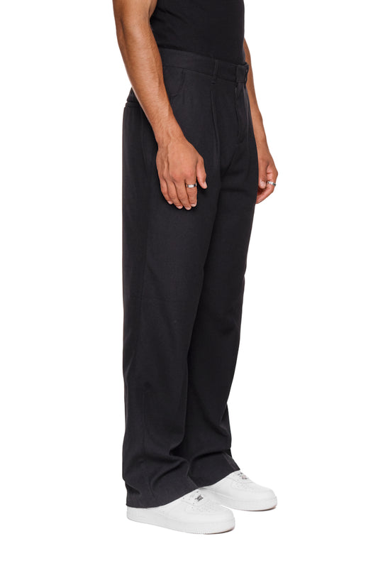 Person trägt CC Dress Pants Regular Black mit französischen Taschen, Bundfalten vorne und gesticktem OLAKALA-Logo über der rechten Gesäßtasche, weiße Schuhe ergänzen den Look.