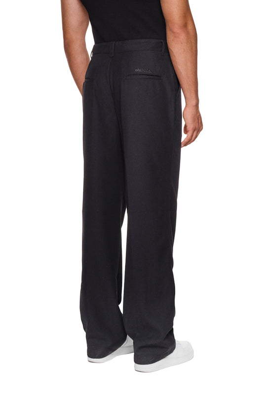 Mann trägt die CC Dress Pants Regular Black von OLAKALA mit französischen Taschen und Paspeltaschen hinten, zeigt asymmetrische Details und gesticktes Logo.