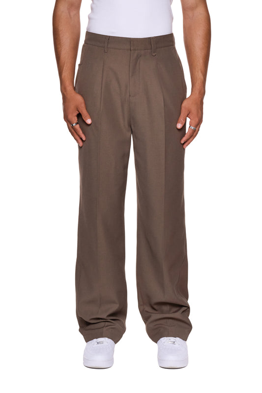 CC Dress Pants Regular Brown mit französischen Taschen, Bundfalten, asymmetrischem Eingriff und gesticktem OLAKALA Logo, getragen von einer Person, inklusive weißem Schuh.