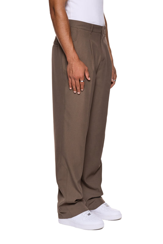 Person trägt braune CC Dress Pants Regular mit französischen Taschen, asymmetrischem Eingriff, Bundfalten und gesticktem OLAKALA-Logo über der rechten Gesäßtasche. Unisex-Design.