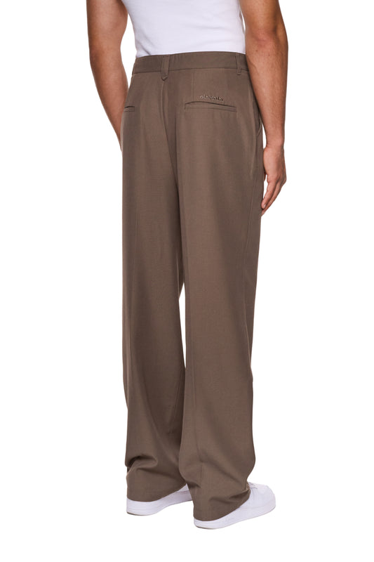 Person trägt CC Dress Pants Regular Brown mit französischen Taschen, asymmetrischem Eingriff und gesticktem OLAKALA-Logo. Paspeltaschen hinten, weiches Köper-Gewebe, reguläre Passform.
