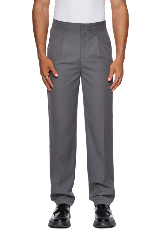 Mann trägt CC Dress Pants Slim Fit Gray mit asymmetrischer Tasche, Bundfalten und gesticktem OLAKALA-Logo über der rechten Gesäßtasche. Schmale Passform, robustes Köper-Gewebe.