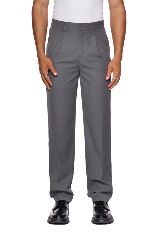 Mann trägt CC Dress Pants Slim Fit Gray mit asymmetrischer Tasche, Bundfalten und gesticktem OLAKALA-Logo über der rechten Gesäßtasche. Schmale Passform, robustes Köper-Gewebe.