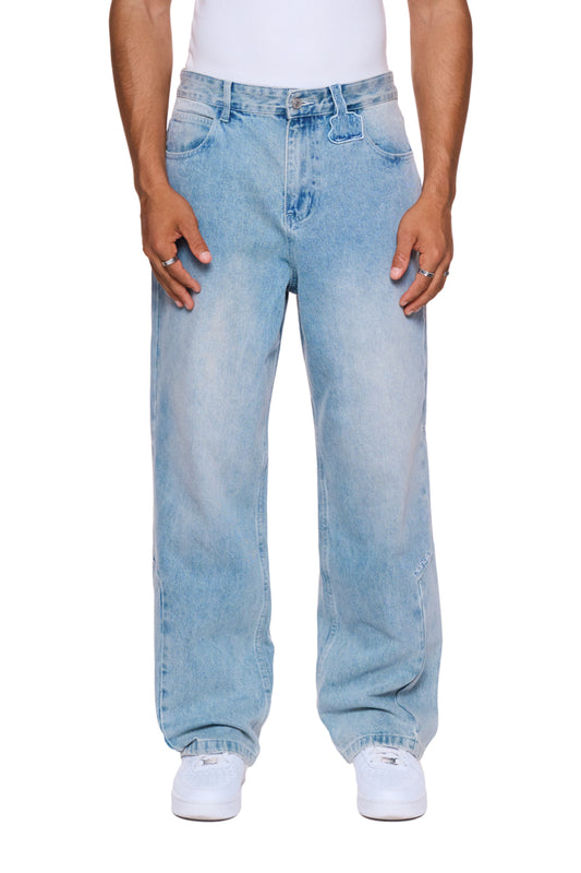 Person trägt CC Denim Pants Regular Blue mit 5-Pocket-Design, versetztem Hosenbund und gesticktem OLAKALA Logo, kombiniert mit weißen Schuhen.