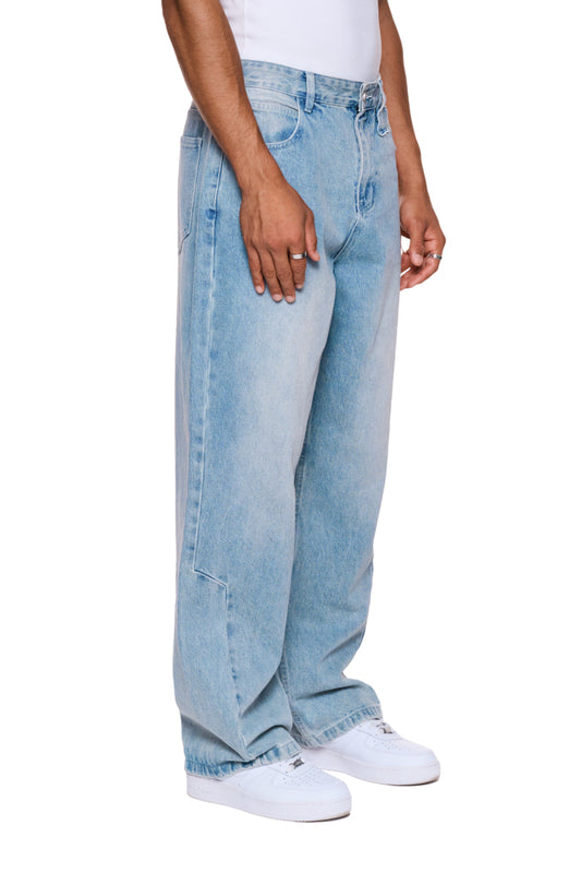 Person trägt CC Denim Pants Regular Blue mit versetztem Hosenbund und gesticktem OLAKALA Logo, betont durch weiße Schuhe.