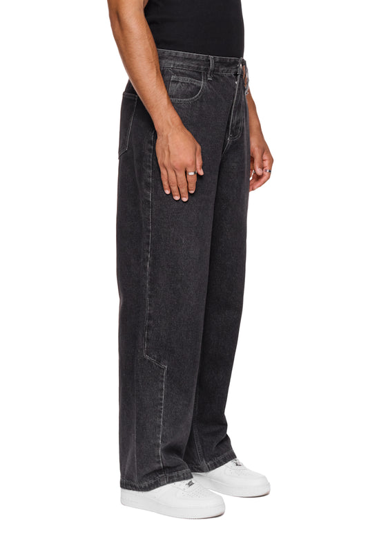 Person trägt CC Denim Pants Regular Black mit versetztem Hosenbund und gesticktem OLAKALA Logo, zeigt seitliche Naht und 5-Pocket-Design.