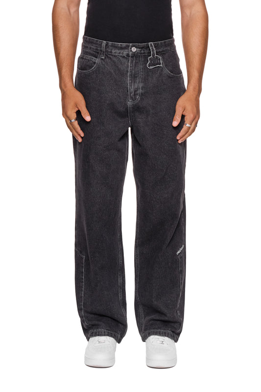 Person trägt CC Denim Pants Regular Black mit versetztem Hosenbund, 5-Pocket-Design und gesticktem OLAKALA-Logo, kombiniert mit weißen Schuhen.