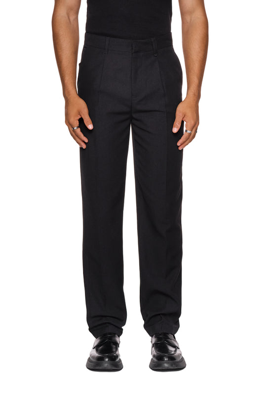 Mann trägt CC Dress Pants Slim Fit Black mit asymmetrischen Taschen und gesticktem OLAKALA Logo, schmale Passform, betont durch Bundfalten und versetzte Seitennaht.
