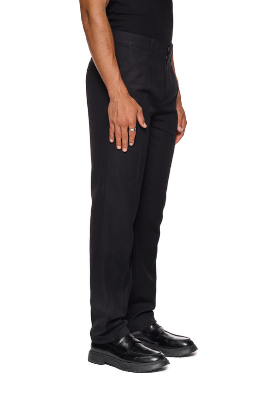Person trägt CC Dress Pants Slim Fit Black, zeigt französische Taschen, asymmetrischen Eingriff, Paspeltaschen hinten, Bundfalten vorne und gesticktes OLAKALA Logo über der rechten Gesäßtasche.