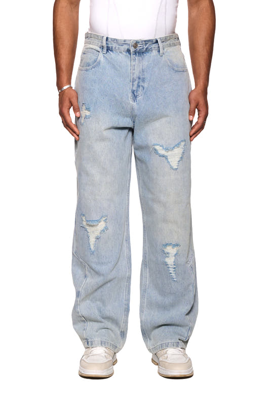 Person trägt Distressed Denim Pants mit versetztem Hosenbund und gesticktem OLAKALA-Logo, sichtbar sind Sneakers und ein Patch an der Gürtelschlaufe.