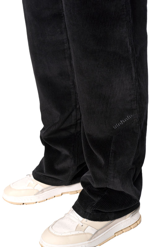 Beine in schwarzen Cordhosen mit asymmetrischer Tasche und gesticktem OLAKALA-Logo, kombiniert mit weißen Schuhen. Unisex, reguläre Passform.