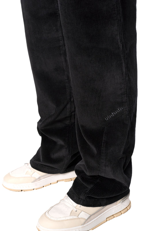 Beine in schwarzen Cordhosen mit asymmetrischer Tasche und gesticktem OLAKALA-Logo, kombiniert mit weißen Schuhen. Unisex, reguläre Passform.
