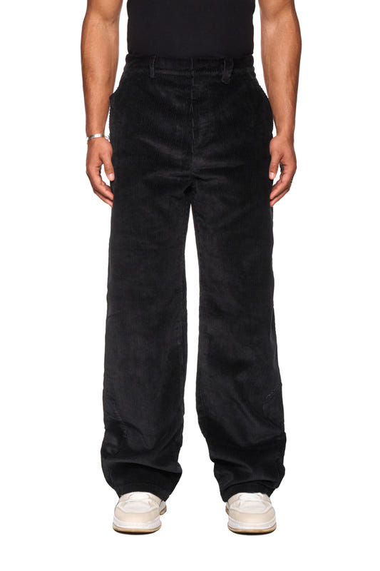Eine Person trägt Corduroy Pants Black mit asymmetrischer Tasche und gesticktem OLAKALA Logo, zeigt stilvolle Details wie Leistentaschen und reguläre Passform.