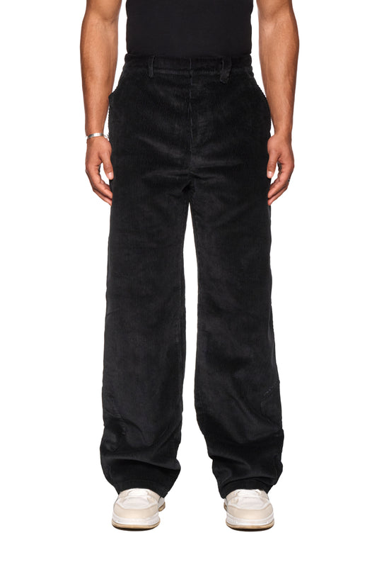 Eine Person trägt Corduroy Pants Black mit asymmetrischer Tasche und gesticktem OLAKALA Logo, zeigt stilvolle Details wie Leistentaschen und reguläre Passform.