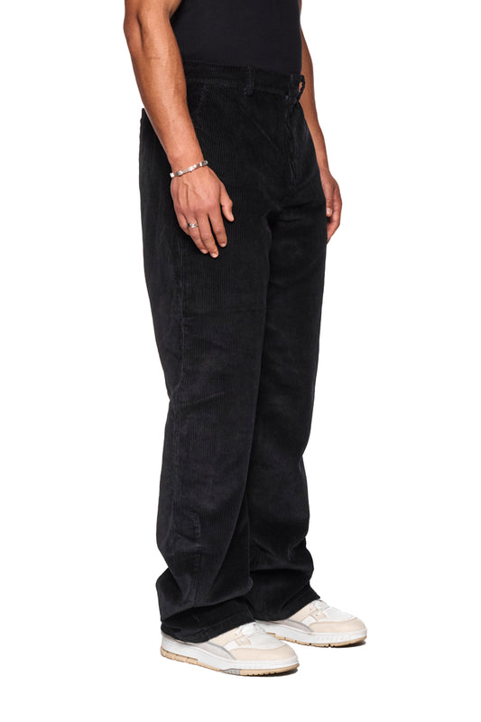 Person trägt schwarze Cordhose mit asymmetrischer Tasche und gesticktem OLAKALA-Logo; Nahaufnahme von Schuhen. Unisex-Design und reguläre Passform.