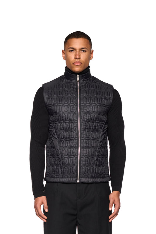 Mann trägt Gilet Vest Black mit Stehkragen und OLAKALA-Logodruck. Sichtbare Reißverschlüsse und Eingriffstaschen betonen das Design.