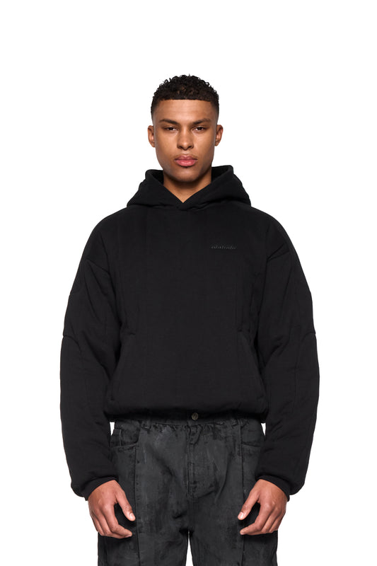 Mann trägt Greyzone Puffer Hoodie mit gesticktem OLAKALA Logo, Ripp-Bündchen und elastischem Saum, in Oversized-Passform. Hände in den Taschen.