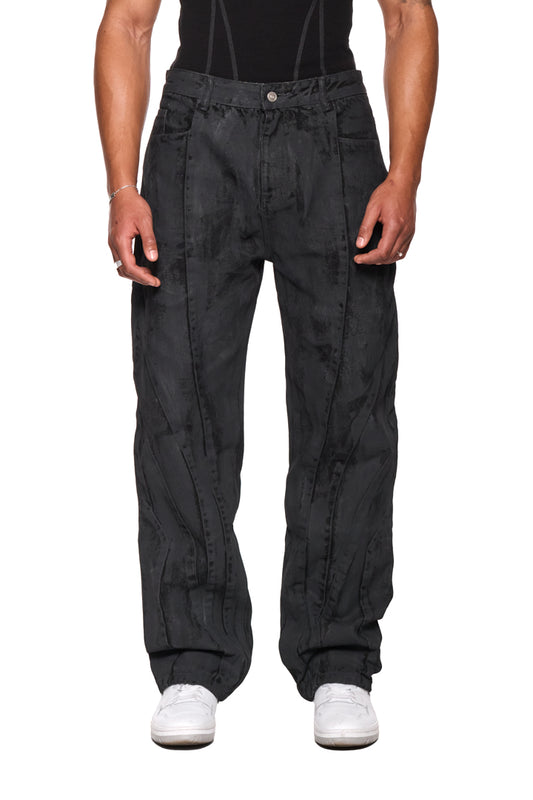 Person trägt CI Coated Denim Pants Regular mit 5-Pocket-Design und Destroyed-Allover-Print, Teilungsnähten und geprägtem OLAKALA Patch, reguläre Passform, unisex.