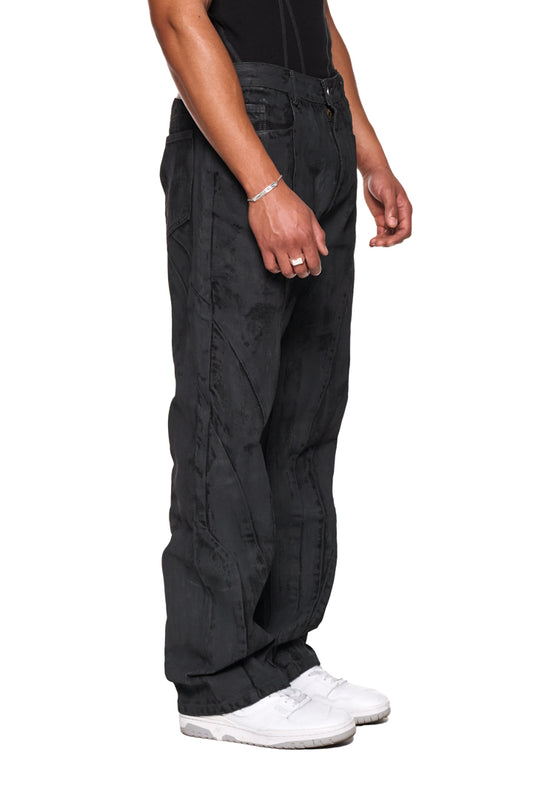 Person trägt OLAKALA CI Coated Denim Pants Regular mit 5-Pocket-Design, Teilungsnähten und versetzter Seitennaht, ergänzt durch ein geprägtes OLAKALA-Patch am Bund.