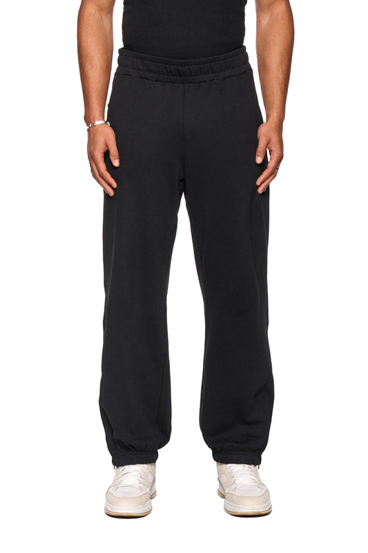 CI Loose Sweatpants Black mit asymmetrischen Taschen und elastischem Bund, getragen von einer Person. Sichtbar sind die Hosenbeine und weiße Schuhe.