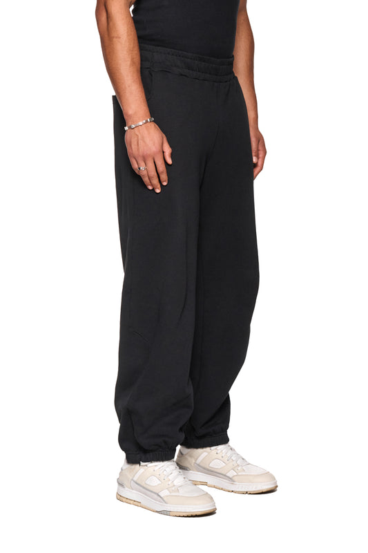 Person trägt CI Loose Sweatpants Black mit asymmetrischer Fronttasche, Kordelzugbund und gesticktem OLAKALA Logo. Weiße Sneaker sind ebenfalls sichtbar.