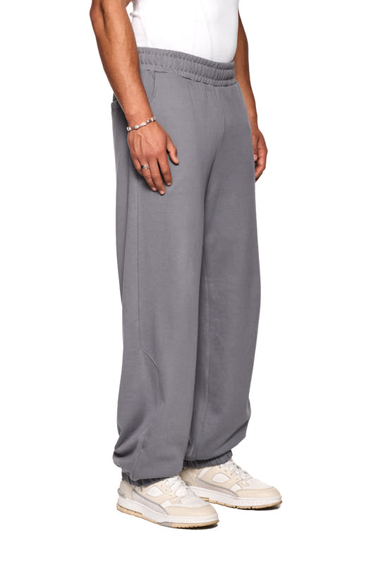 Person trägt CI Loose Sweatpants Gray mit asymmetrischen Taschen, elastischem Bund und gesticktem OLAKALA Logo.