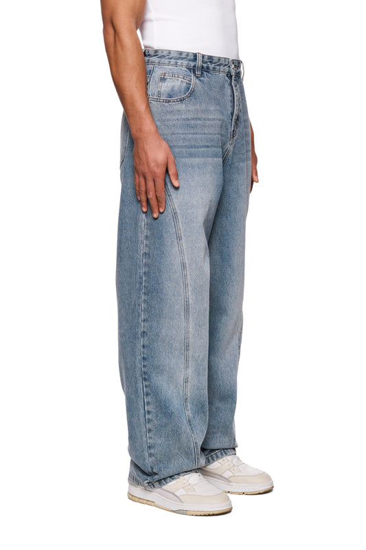 Person trägt CC Core Denim Pants Blue mit 5-Pocket-Style, robuster Denim, lockere Passform, weiße Schuhe sichtbar, Hand in Tasche.