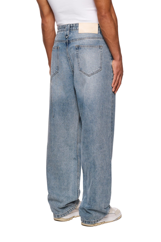 Person trägt CC Core Denim Pants Blue aus robustem, verwaschenem Denim im 5-Pocket-Style mit OLAKALA PU-Patch und lockerer Passform.