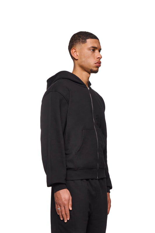Mann im CC Core Zip Hoodie Schwarz mit Kängurutasche und Rippbündchen, zeigt Detail des Reißverschlusses. Perfekt für Komfort und Langlebigkeit.