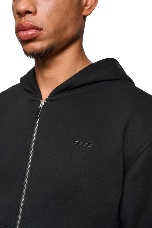 Mann trägt CC Core Zip Hoodie Black mit Reißverschluss und Kängurutasche, bietet Langlebigkeit und Komfort durch strapazierfähige Mischfaser und verlegte Nähte.
