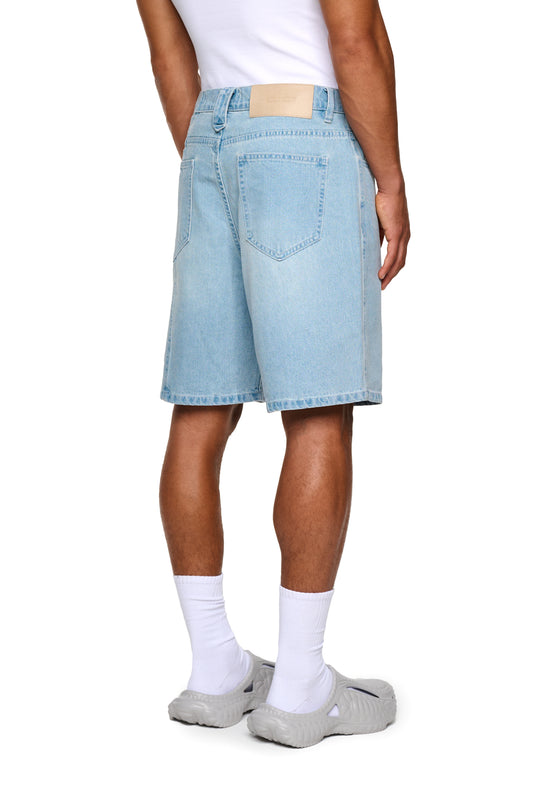 Nahaufnahme von Beinen in weißen Socken, die 0asis Denim Shorts Blue tragen, mit geprägtem OLAKALA-Patch und Logo-Stickerei sichtbar.