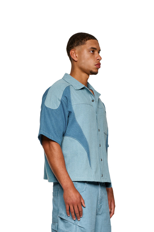 Nahaufnahme eines OLAKALA®-Echoes Denim Icon Shirt Blue mit kubanischem Kragen und Brusttasche mit Logo-Stickerei, präsentiert auf einem Modell, Fokus auf Ärmel und Schulter.