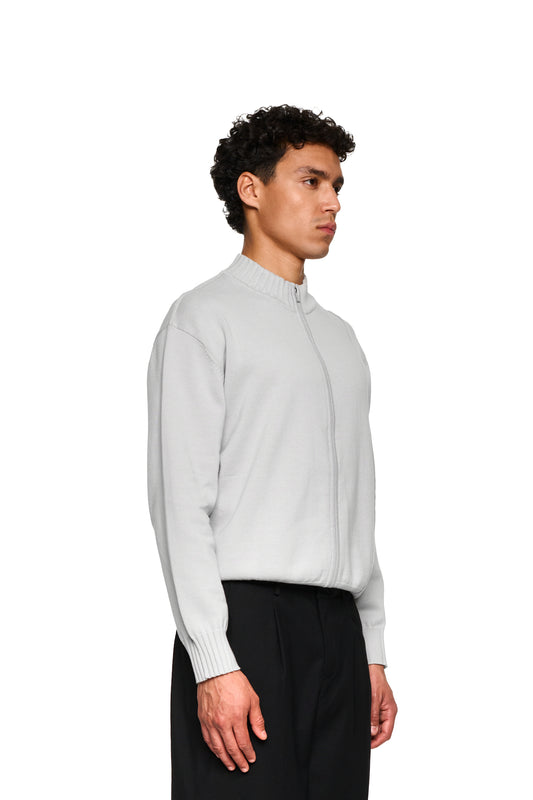 Hand in weißem Pullover zeigt die Sommer-Strickjacke Light Gray mit asymmetrischer Vorderseite und Metallreißverschluss, ideal für warme Abende, von OLAKALA.