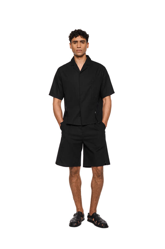 Schwarze Leinen-Shorts mit Bundfalten, französischen Taschen, asymmetrischem Eingriff, rückseitigen Leistentaschen und seitlichen Schlitzen, aus leichtem, atmungsaktivem Gewebe. Unisex, reguläre Passform.