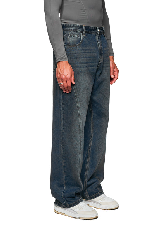 Person trägt CC Core Denim Pants Tinted Indio mit lockerer Passform und versetzter Seitennaht, kombiniert mit weißen Sneakers. Dezente, moderne Interpretation klassischer Jeans.