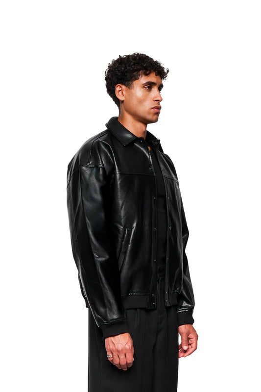 Junger Mann im Profil trägt CC Leather Jacket Icon Black mit Druckknopf-Leistentaschen, gesticktem OLAKALA-Icon, Oversized-Passform, veganem Lederimitat und Reißverschluss-Innentasche.