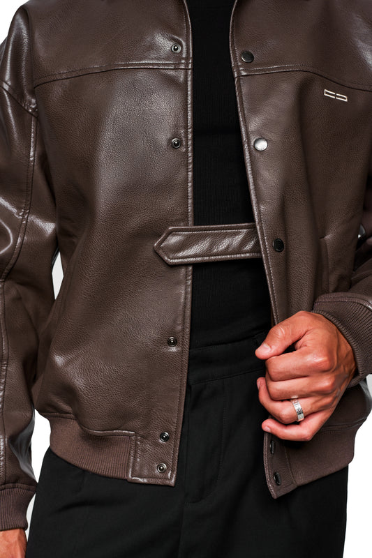 Person trägt CC Leather Jacket Icon Brown aus veganem Lederimitat mit Druckknopfdetails und gesticktem OLAKALA-Icon, kombiniert mit schwarzem Shirt und Hose.