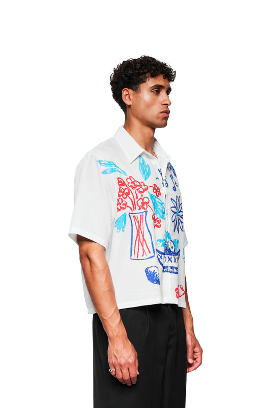 Junger Mann im Profil trägt das Crayon Summer Shirt Graphic mit skizzenhaften Motiven und kubanischem Kragen, kombiniert mit schwarzen Hosen. Perfekt für heiße Sommertage.