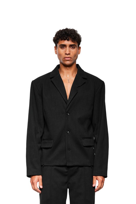Männliches Model trägt Exquisite Blazer Black mit asymmetrischem Revers, fake-Taschen und herausnehmbaren Schulterpolstern. Unisex-Design, schlanke Passform, minimalistischer Stil.