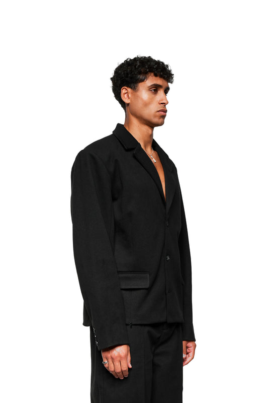 Männliches Modell trägt Exquisite Blazer Black mit asymmetrischem Revers, Zweinahtärmel, und herausnehmbaren Schulterpolstern, kombiniert mit schmal geschnittenen schwarzen Hosen.