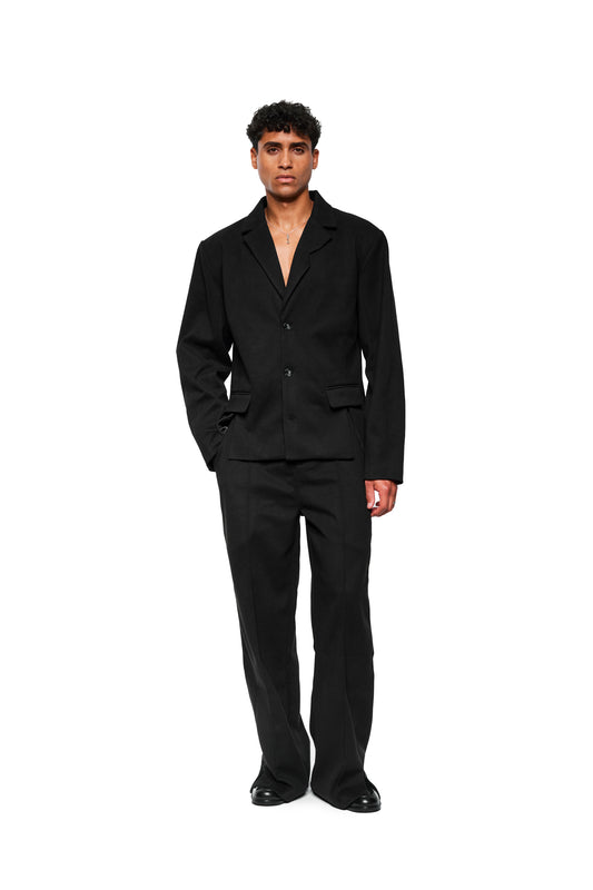 Male model in Exquisite Blazer Black, one-piece, with asymmetrical lapels, two-seam sleeves and walking slit. Combined with matching trousers and black shoes.