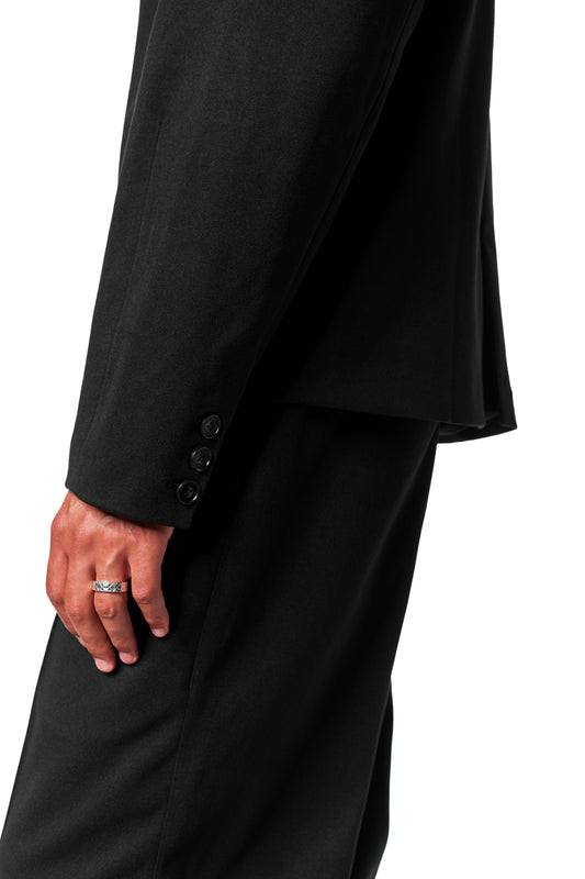 Person wearing Exquisite Blazer Black with asymmetrical lapel, three buttons on the cuff and a silver ring on the finger. Tight fit, unisex design.