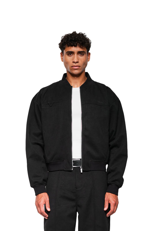 Junger Mann trägt Exquisite Bomber Jacket Black mit geripptem Stehkragen, Brusttasche, Reißverschluss-Innentasche und gesticktem OLAKALA-Icon, präsentiert in minimalistischer, unisex Passform.