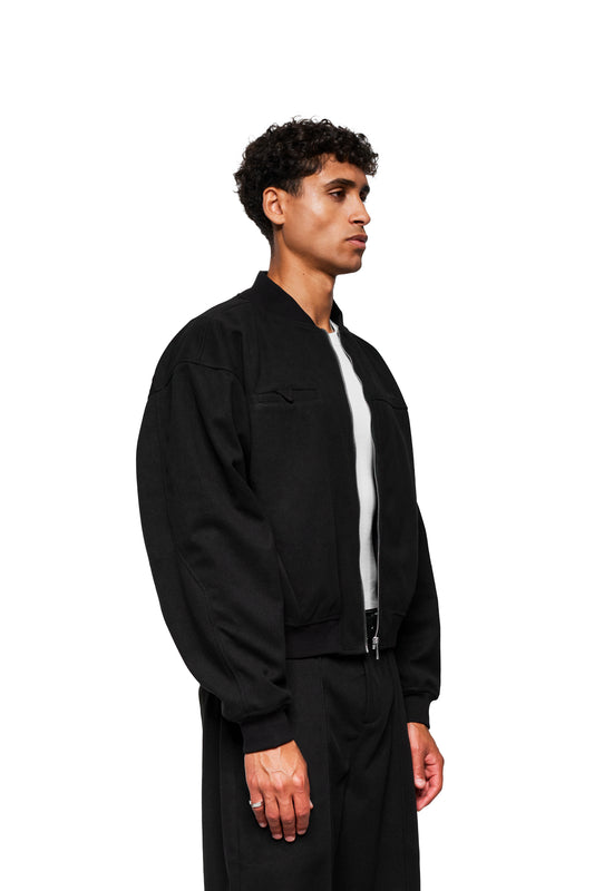 Junger Mann im Profil trägt Exquisite Bomber Jacket Black mit Stehkragen, Brusttasche und gesticktem OLAKALA-Icon, dazu passende schwarze, locker geschnittene Hose.
