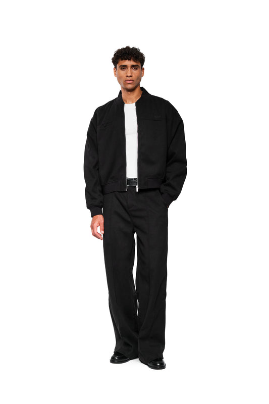 Male model in Exquisite Dress Pants Black with a wide fit, piping at the front and embroidered OLAKALA icon, combined with a black jacket and white T-shirt.