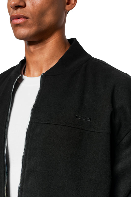 Man wearing Exquisite Bomber Jacket Black with ribbed stand-up collar and embroidered OLAKALA icon. Shows chest pocket and slit pockets worn over a white T-shirt.