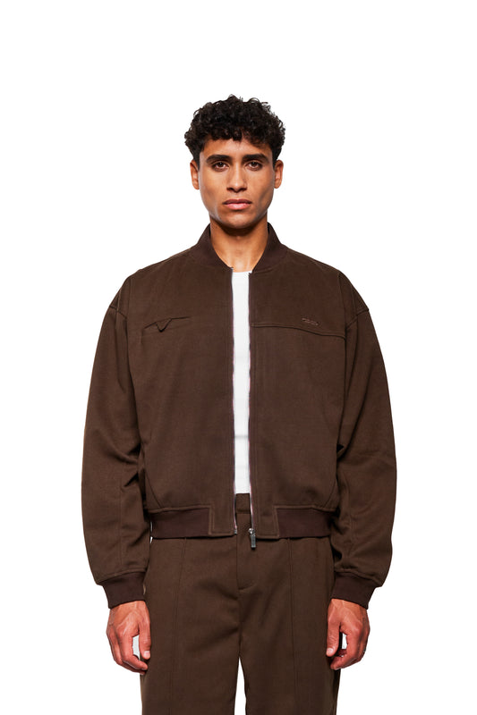 Modell trägt Exquisite Bomber Jacket Brown mit geripptem Stehkragen, Brusttasche und gesticktem OLAKALA Icon, kombiniert mit passenden Hosen, präsentiert auf weißem Hintergrund.