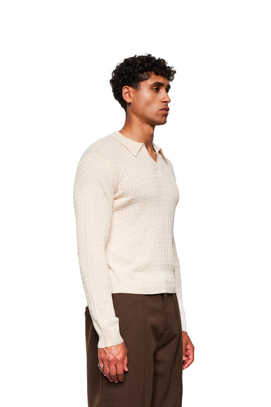 Männliches Model im Exquisite Cable Knit Polo Creme, mit Zopf-Strickmuster, V-Ausschnitt und eng anliegender Passform, kombiniert mit hochgeschnittenen braunen Hosen.