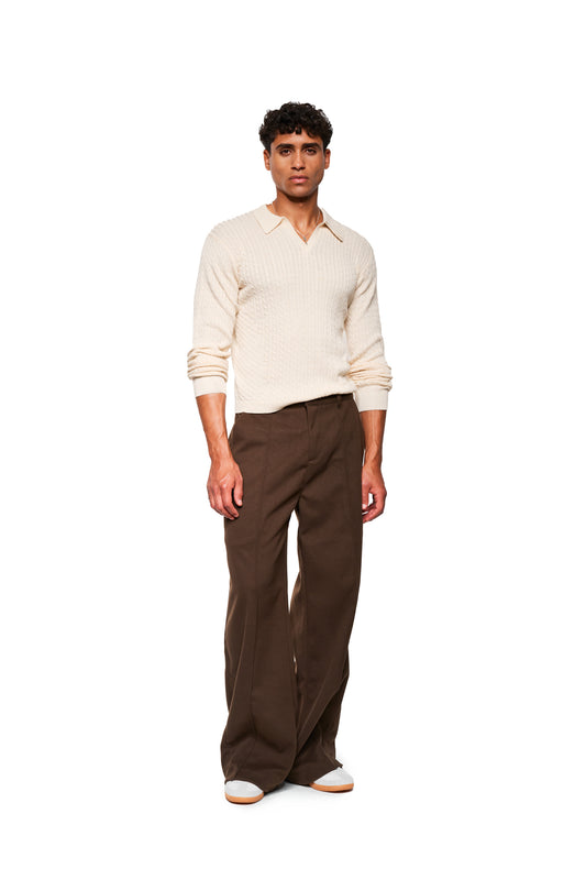 Male model wears Exquisite Cable Knit Polo Cream, a cream-colored, cable-knit V-neck sweater with a small collar, combined with high-waisted, wide brown pants and white sneakers.